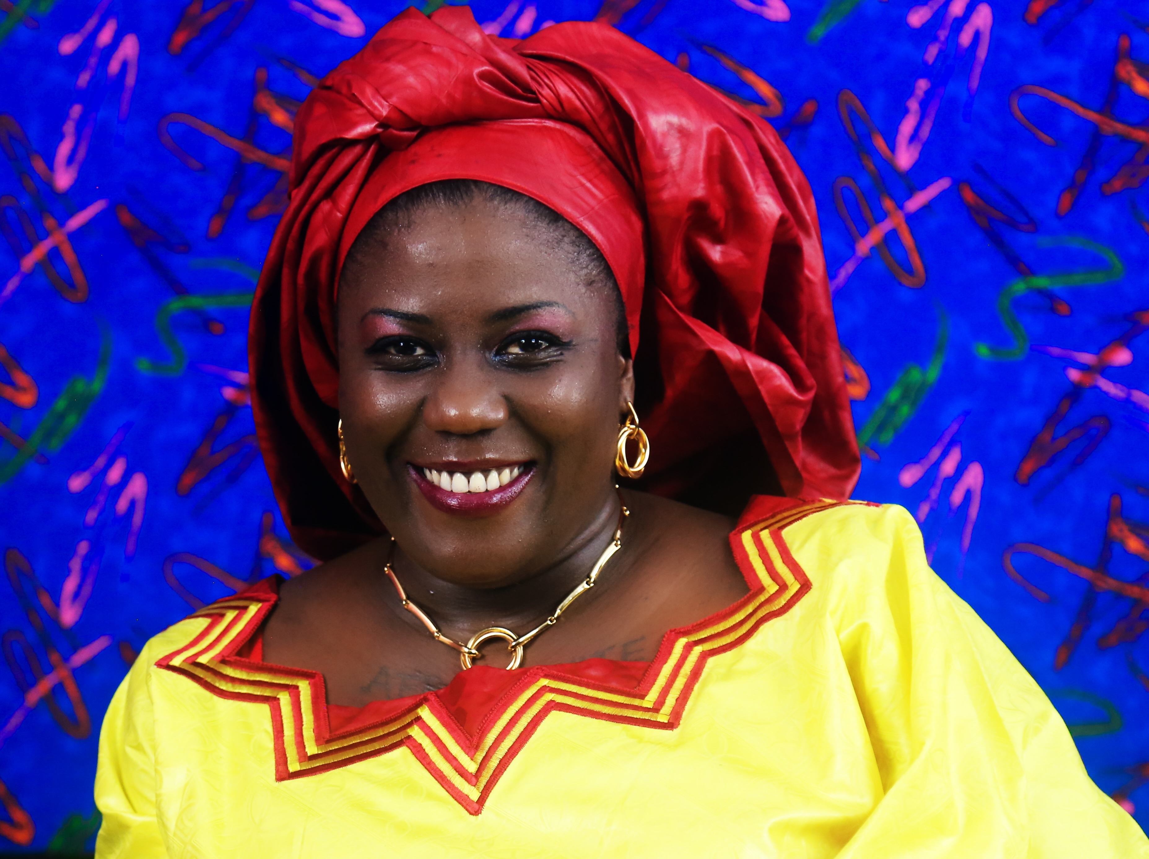 Maidem Kayem, CFO and Talent Acquisition, smiling woman wearing yellow top with red trim and red head wrap against blue patterned background